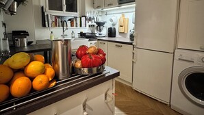 A modern kitchen with white cabinets, a stainless steel sink, and a built-in oven. There are oranges and tomatoes on the counter, and a microwave above the stove.