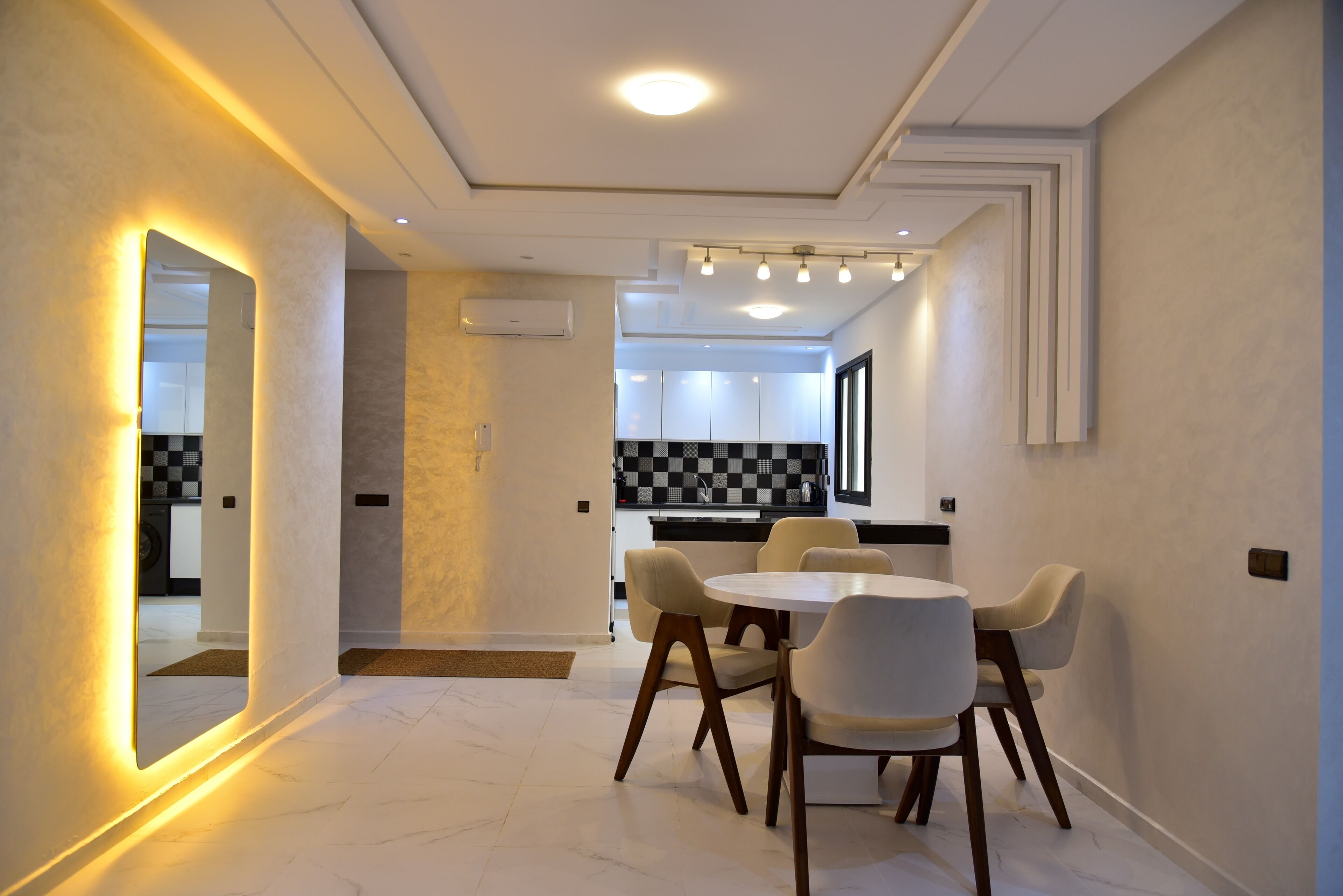 Un'area pranzo moderna con un tavolo rotondo e sei sedie, una cucina sullo sfondo e un corridoio con uno specchio e illuminazione a incasso.