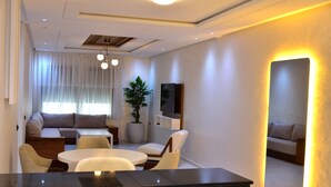 A modern living room with a sofa set, a dining table, and chairs. There is a large mirror on the wall and a potted plant.