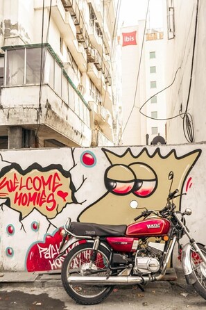 A motorcycle parked in front of a wall with graffiti, including a cartoon face and the words "WELCOME HOMIES". - The Pink Chapter Hostel, Jaipur (Jaipur)