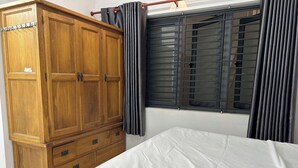 A bedroom with a wooden wardrobe, a bed with white bedding, and a window with vertical blinds. - T&M Homestay (Ho Chi Minh City)