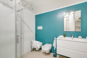 A modern bathroom with a glass shower enclosure, a bidet, a white sink with a mirror, and a teal accent wall.