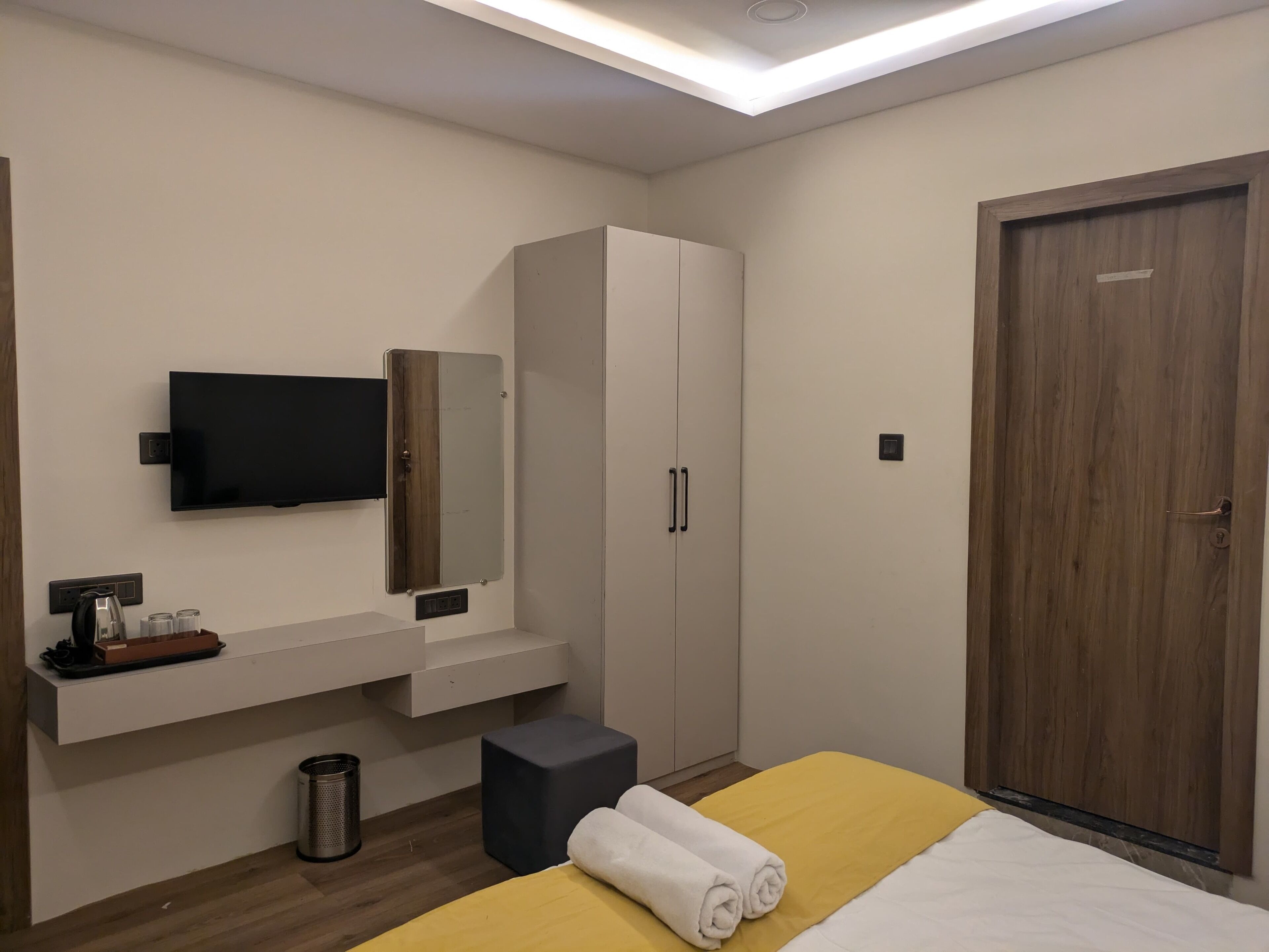 A modern hotel room with a bed, a TV mounted on the wall, a wardrobe, and a wooden door.
