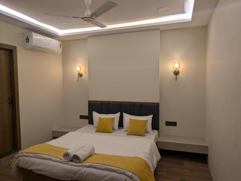 A hotel room with a bed, ceiling fan, wall-mounted air conditioner, and two wall sconces.