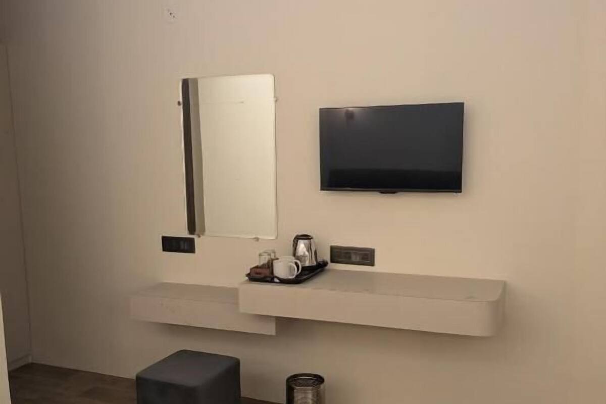 A modern hotel room with a bed, a flat-screen TV mounted on the wall, a small bedside table with a kettle and cups, a trash can, and a grey ottoman.