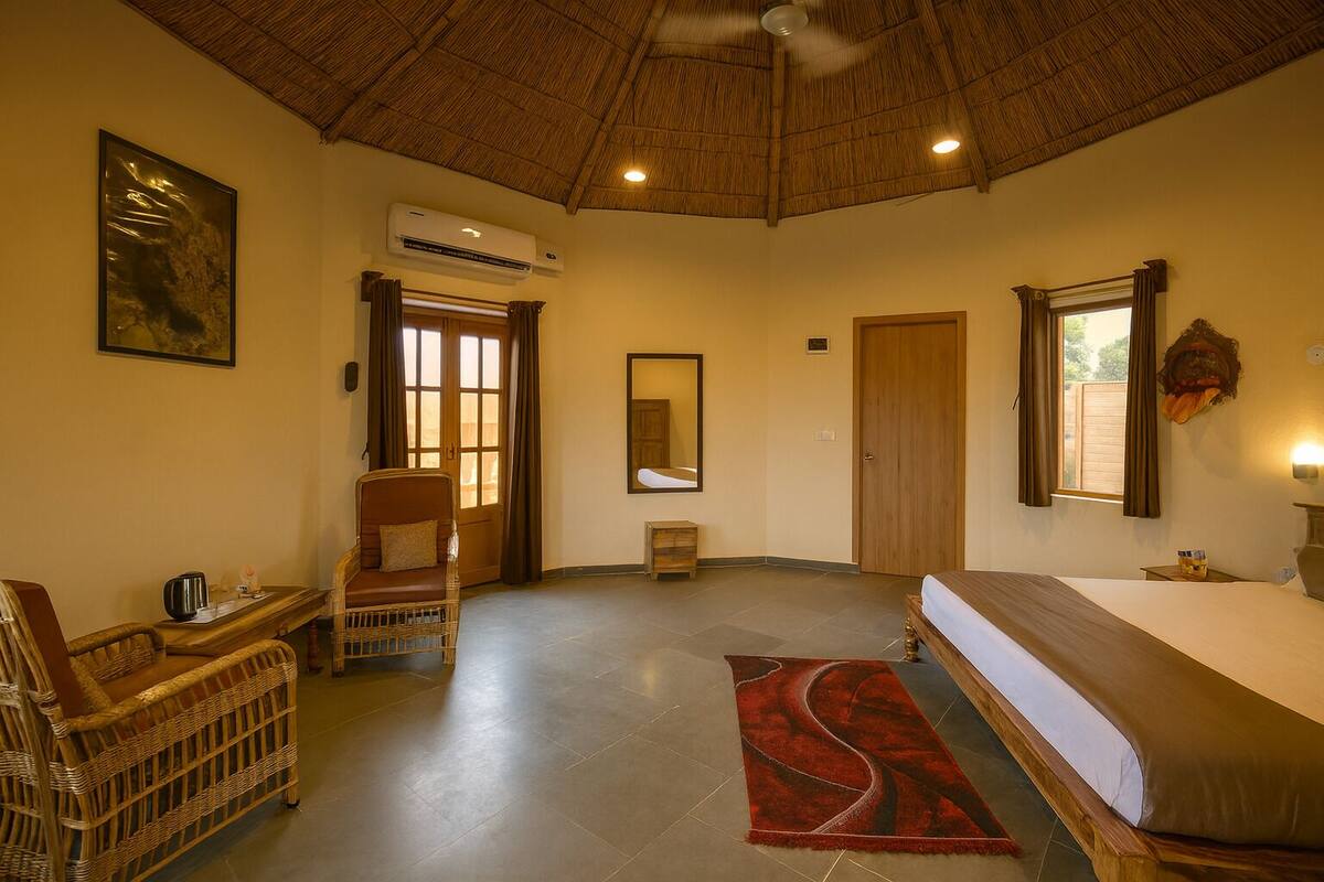 A hotel room with a bed, a chair, a small table, an air conditioning unit, a mirror, and a window with curtains.