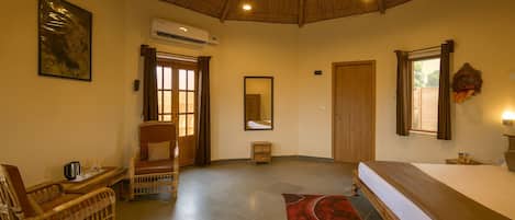 A hotel room with a bed, a chair, a small table, an air conditioning unit, a mirror, and a window with curtains.