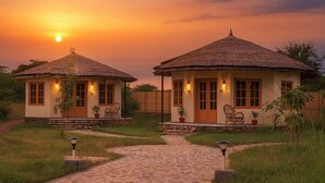 Two thatched-roof cottages with stone foundations, each with a porch and seating area, set against a sunset backdrop. - Jawai Vanya Bagh - Luxury Leopard Safari Resort (Bali)