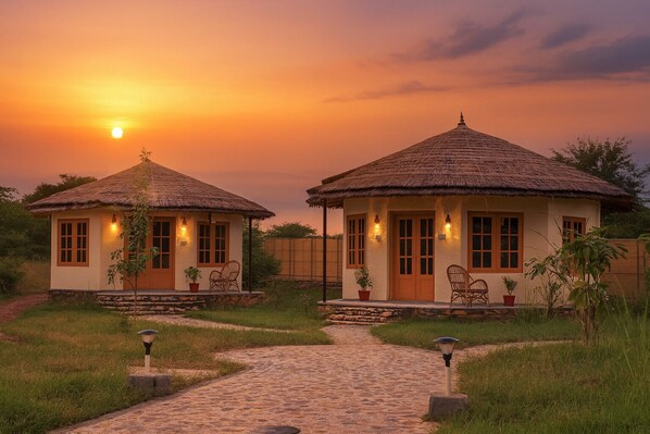 Two thatched-roof cottages with stone foundations, each with a porch and seating area, set against a sunset backdrop.