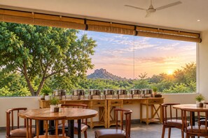 A dining area with round wooden tables and chairs, a buffet setup with metal serving dishes, and a large window offering a view of trees and a mountain. - Jawai Vanya Bagh - Luxury Leopard Safari Resort (Bali)