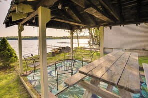 Un espacio al aire libre techado con una mesa de picnic de madera y sillas de metal, con vista a un lago.