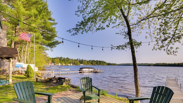 Une terrasse en bois avec deux fauteuils Adirondack verts donnant sur un lac, une jetée et un petit bateau amarré.