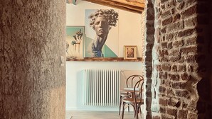 Een smalle gang met openliggende bakstenen muren, een houten plafond en een radiator. Er hangt een lijst met een afbeelding aan de muur en er staat een stoel in de hoek.