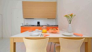 A modern kitchen with orange cabinets, a white countertop, and a dining area with two chairs and a table set for breakfast. - Holiday apartment with air conditioning, balcony and pool (Lignano Sabbiadoro)