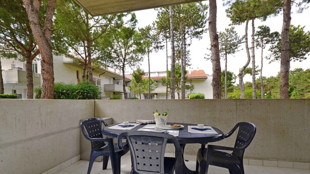 An outdoor patio with a round table and four chairs, surrounded by a low wall and trees. - Holiday apartment with air conditioning, balcony and pool (Lignano Sabbiadoro)