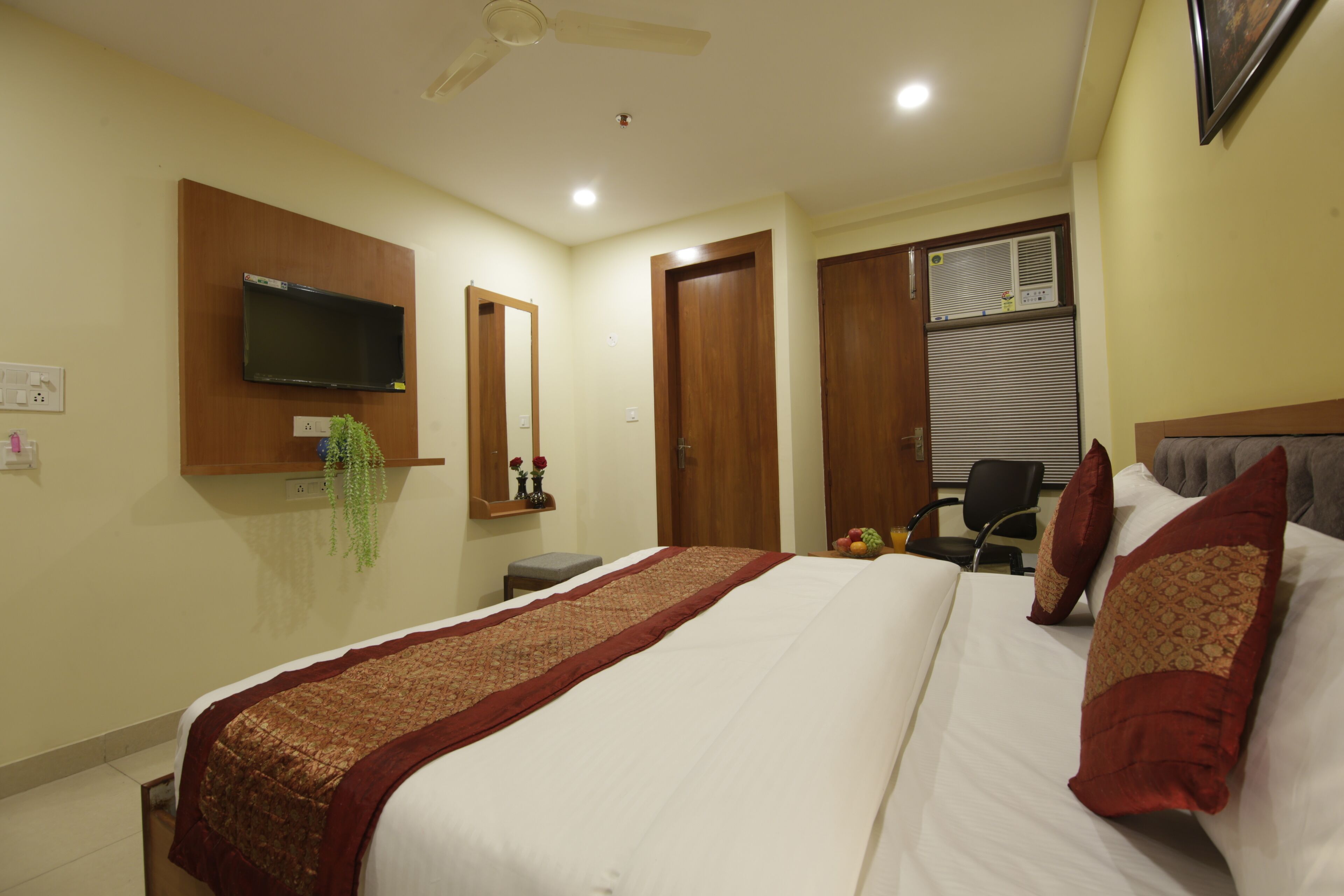 A hotel room with a bed, a television mounted on the wall, a ceiling fan, and a window with blinds.