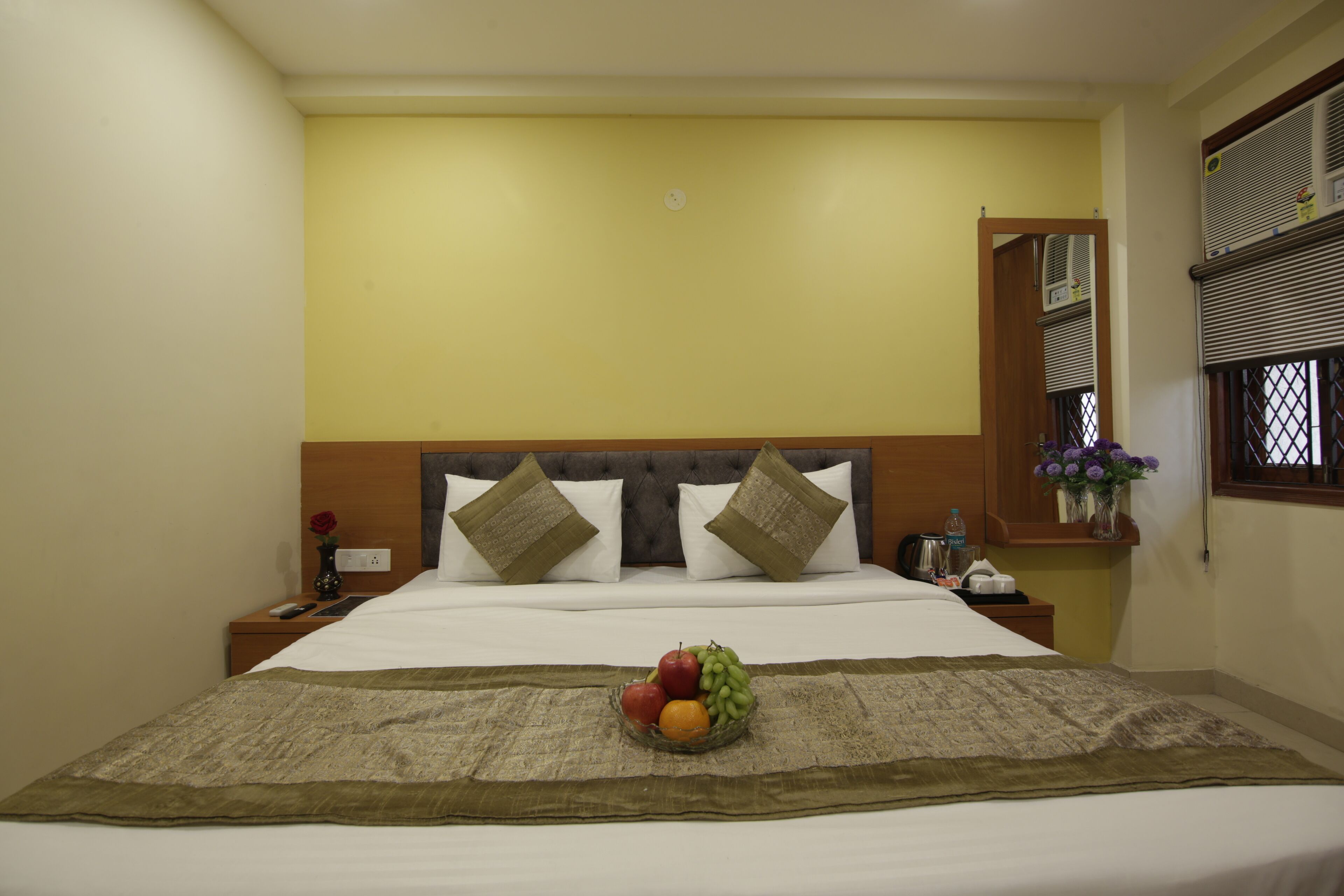 A hotel room with a bed, a bedside table with a vase of flowers, and a window with blinds.