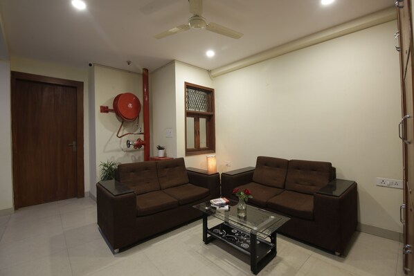 A living room with two brown sofas, a glass coffee table, a ceiling fan, and a red water heater on the wall. - Hotel TMS Grand (New Delhi)
