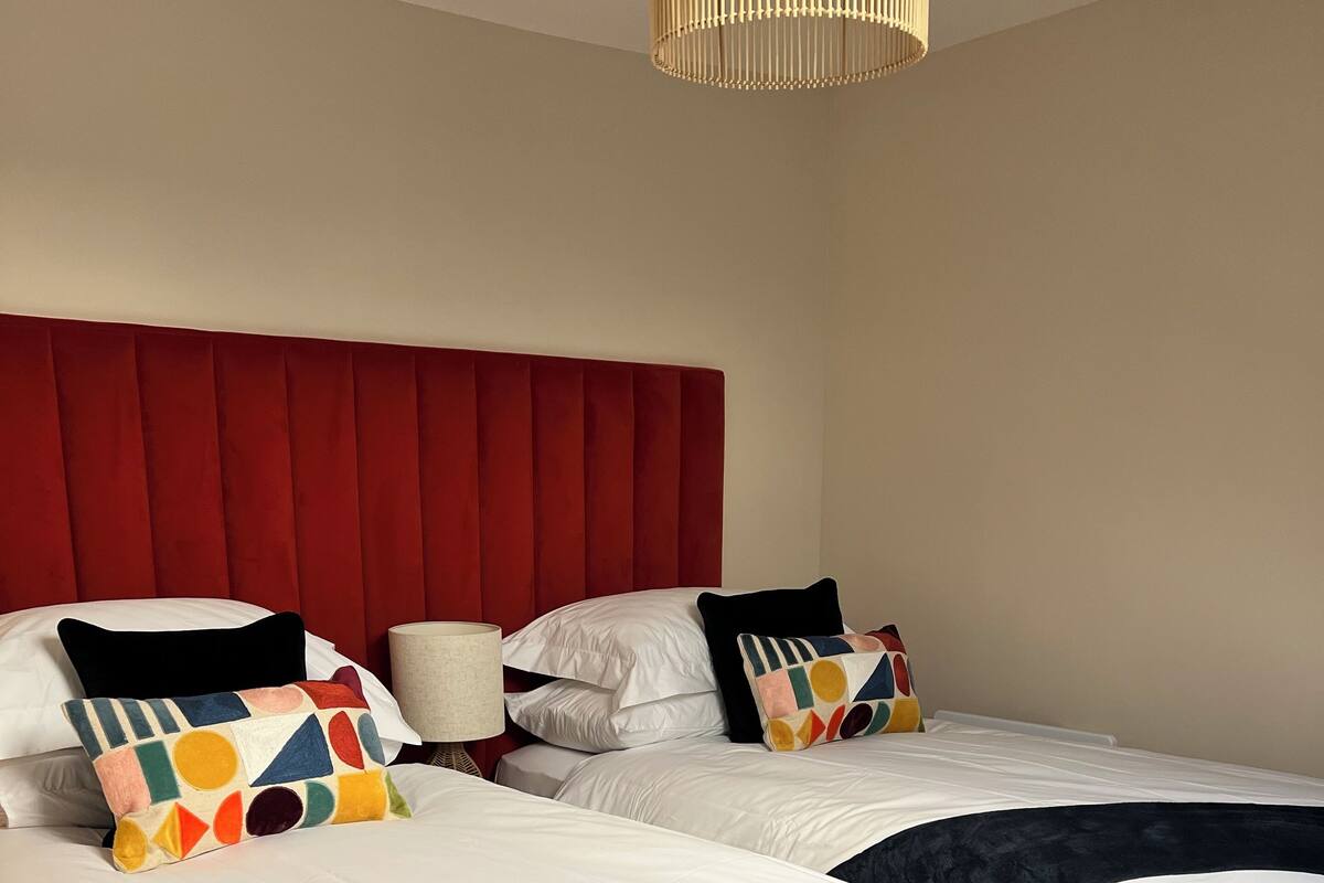 A hotel room with two beds, each with white linens and navy blue throw blankets. The headboard is red, and there is a lamp on the nightstand.