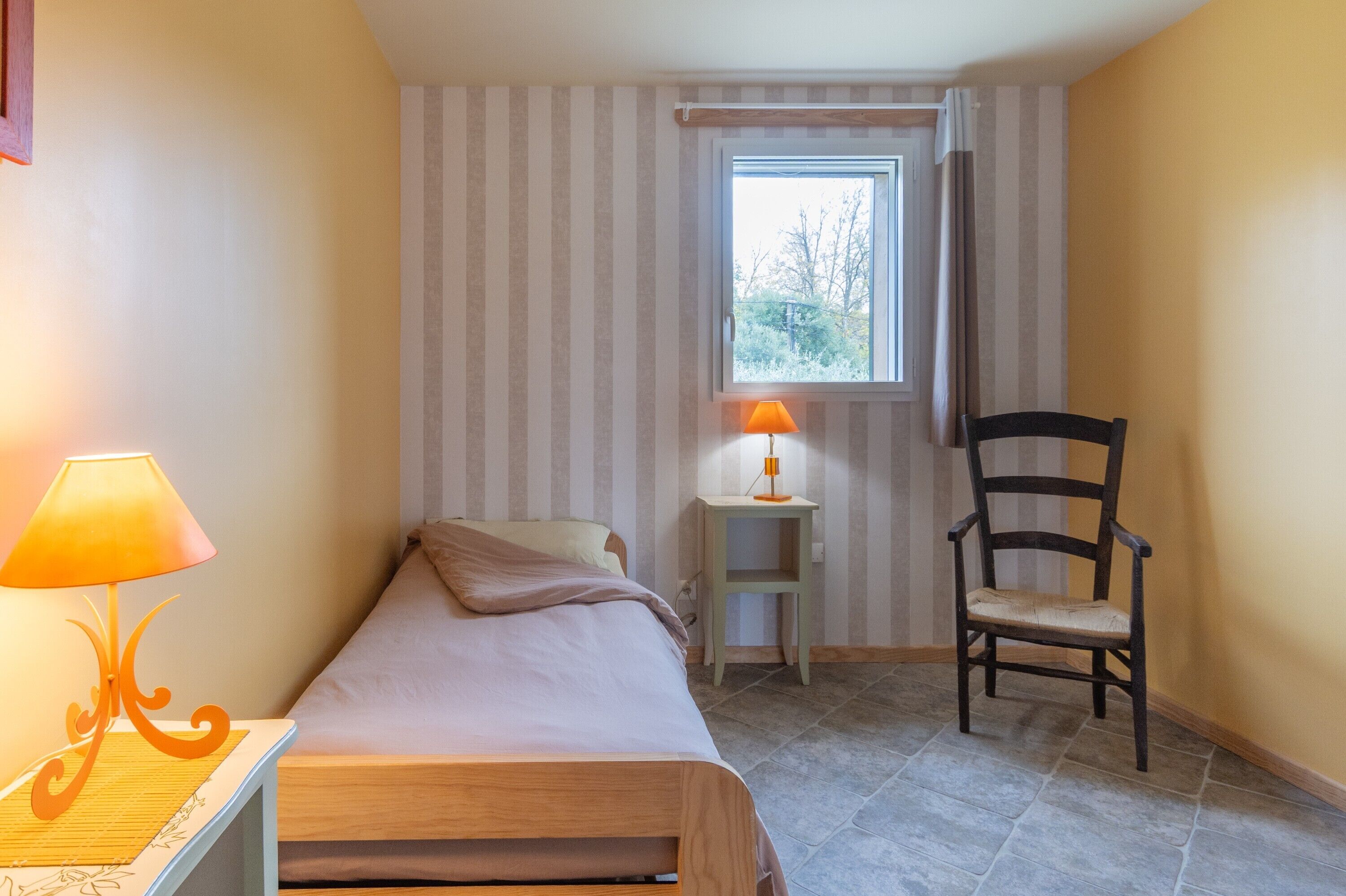 Een kleine kamer met een bed, een stoel, een nachtkastje met een lamp en een raam met gordijnen.