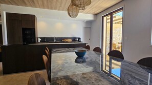 A modern kitchen with dark cabinetry, a central island, and a dining area with a marble table and chairs. - VILLA MODERNE EN CORSE - Sea and mountain views, swimming pool, pool house (CALENZANA)
