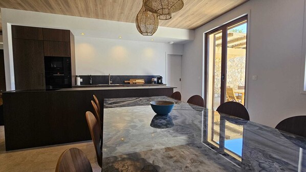 Una cocina moderna con armarios oscuros, una isla central y una zona de comedor con una mesa y sillas de mármol.