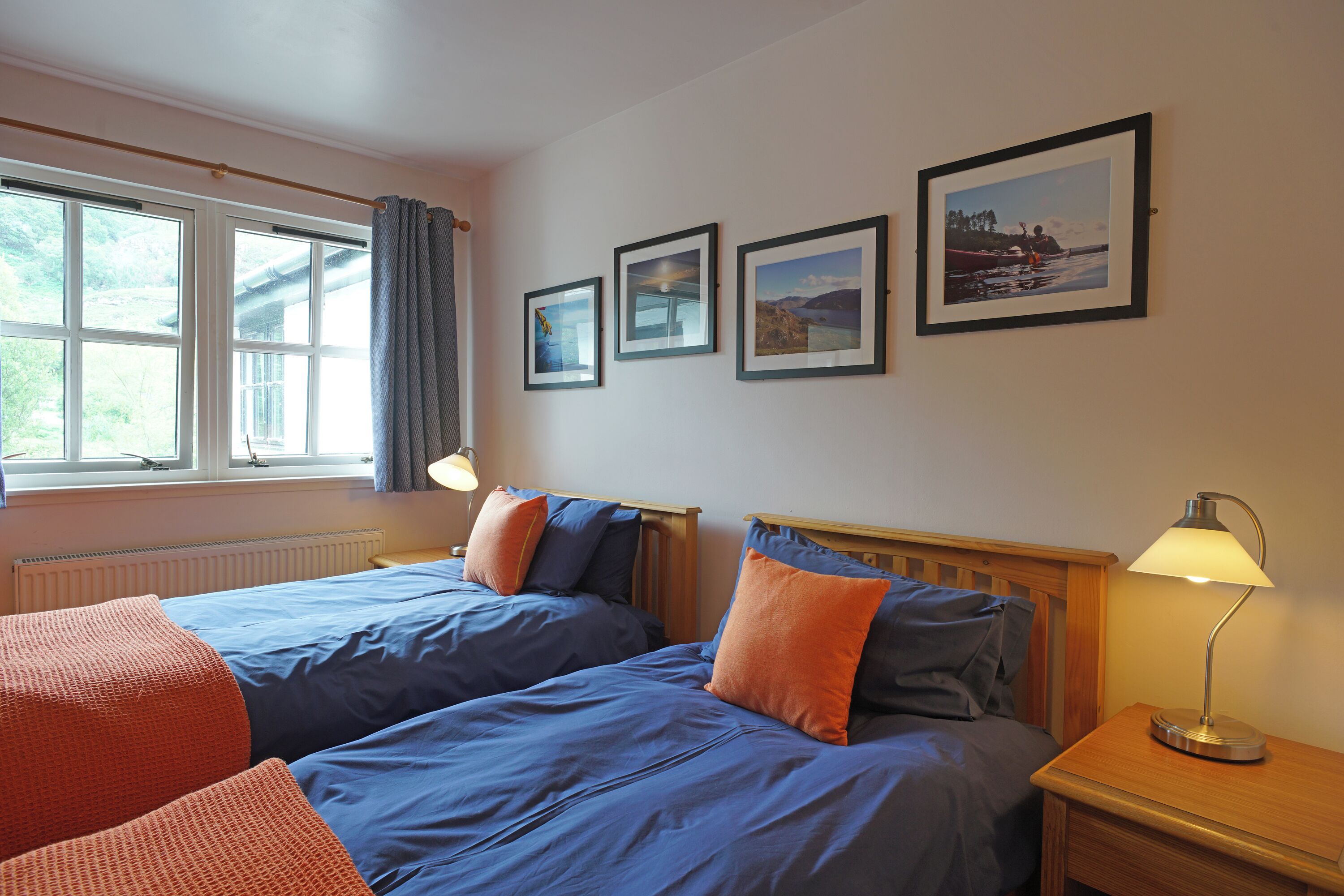 A hotel room with two beds, a nightstand with a lamp, a window with curtains, and three framed pictures on the wall.