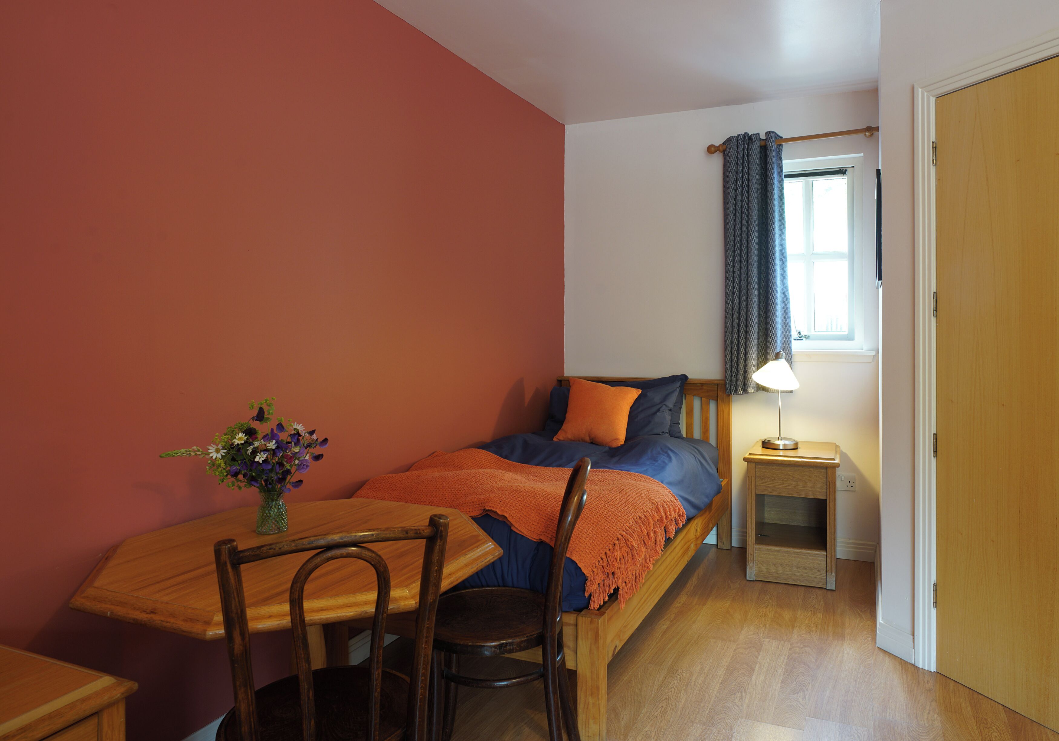 Une chambre à coucher avec un lit, une table de chevet, une lampe, une chaise et une table sur laquelle est posé un vase de fleurs.