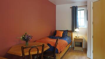 Une chambre à coucher avec un lit, une table de chevet, une lampe, une chaise et une table sur laquelle est posé un vase de fleurs.