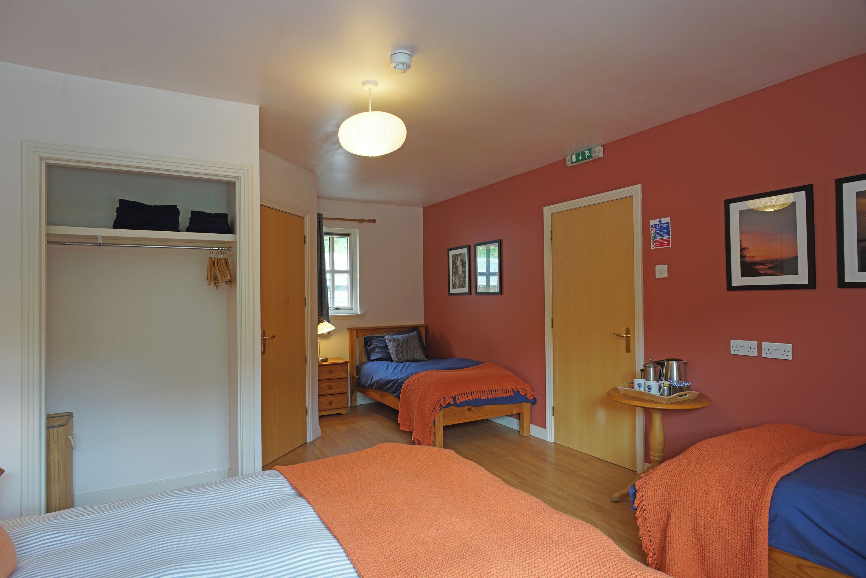 A bedroom with two beds, a wardrobe, a bedside table with a lamp, and a wall-mounted shelf.