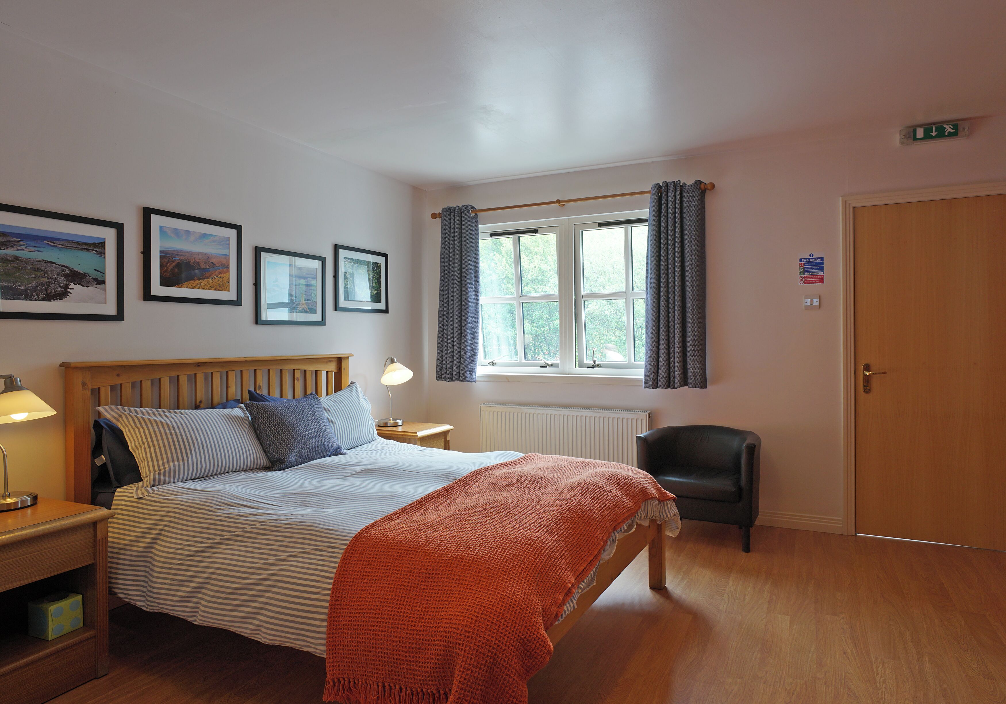 Une chambre à coucher comprenant un lit, une table de chevet, une lampe, une chaise, une fenêtre avec des rideaux et des tableaux encadrés accrochés au mur.
