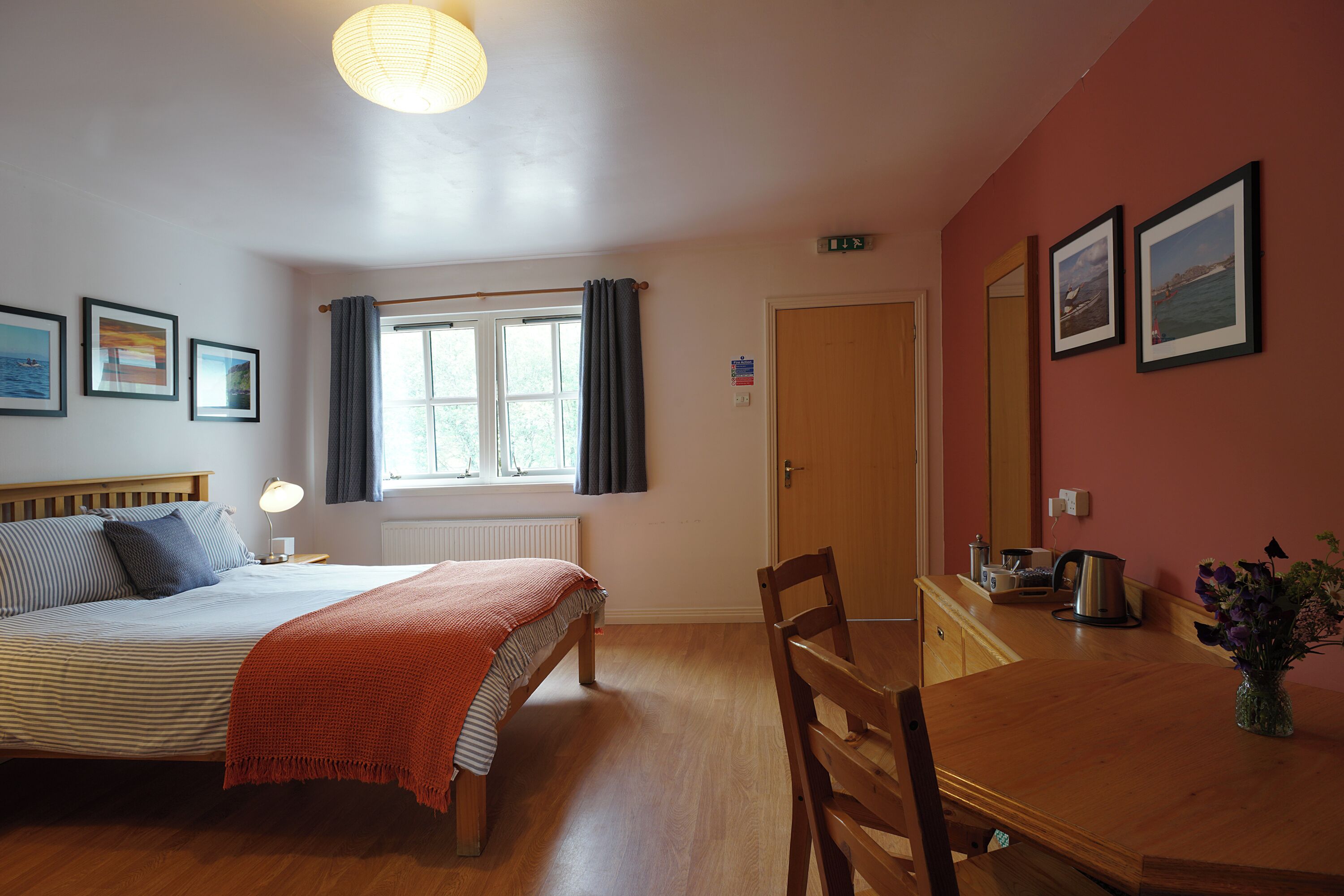 A bedroom with a bed, bedside table, lamp, window with curtains, and a wooden dresser with a mirror.
