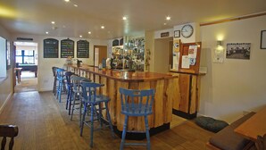 A bar area with wooden bar counter and blue bar stools. There are signs on the wall and a clock. - Glenuig Inn (Lochailort)