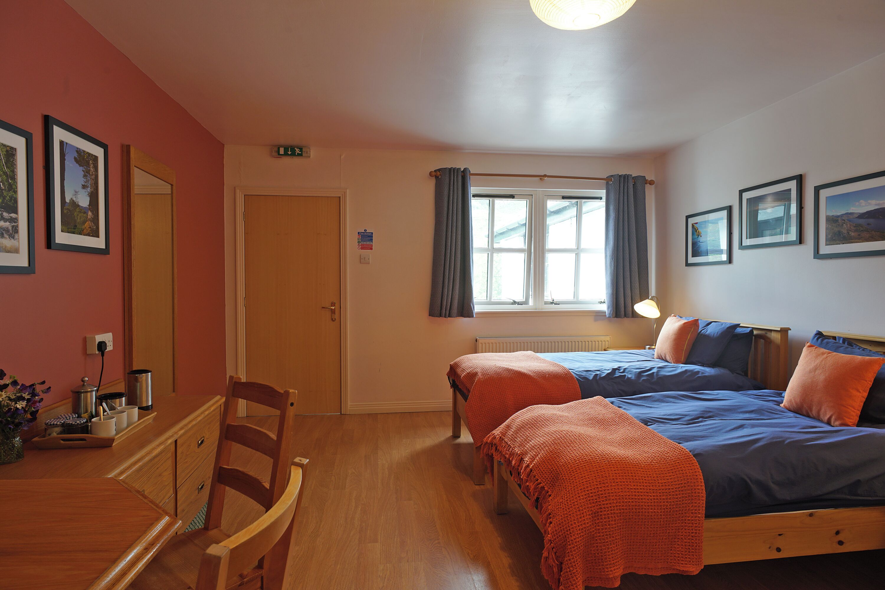 Una habitación con dos camas, una mesa de madera, sillas, una ventana con cortinas y cuadros en la pared.