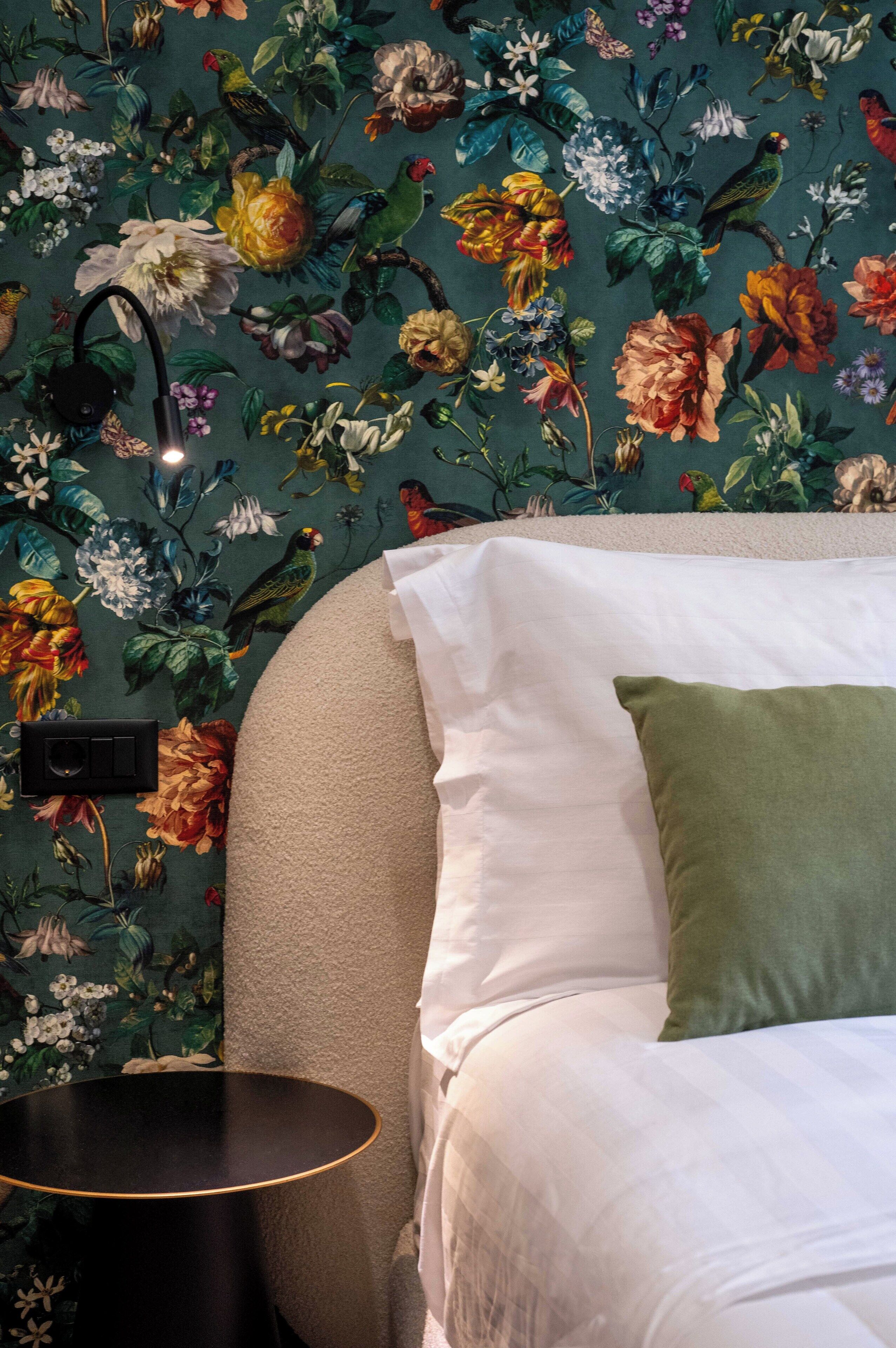 A bedroom with a floral wallpaper, a round black side table, and a bed with white and green pillows.