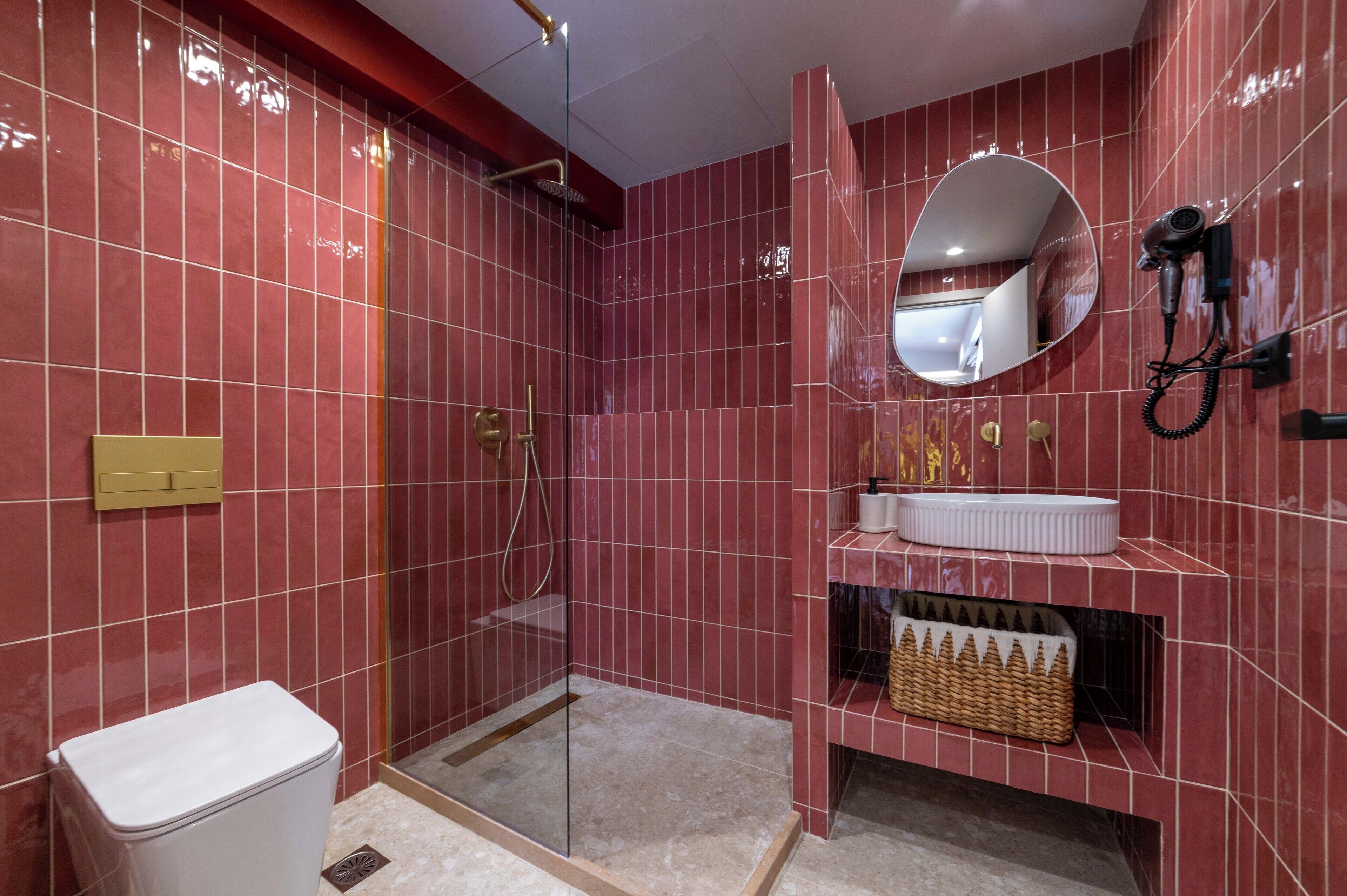 Un baño moderno con baldosas rojas, cabina de ducha con paredes de vidrio, lavabo blanco y secador de pelo fijado en la pared.