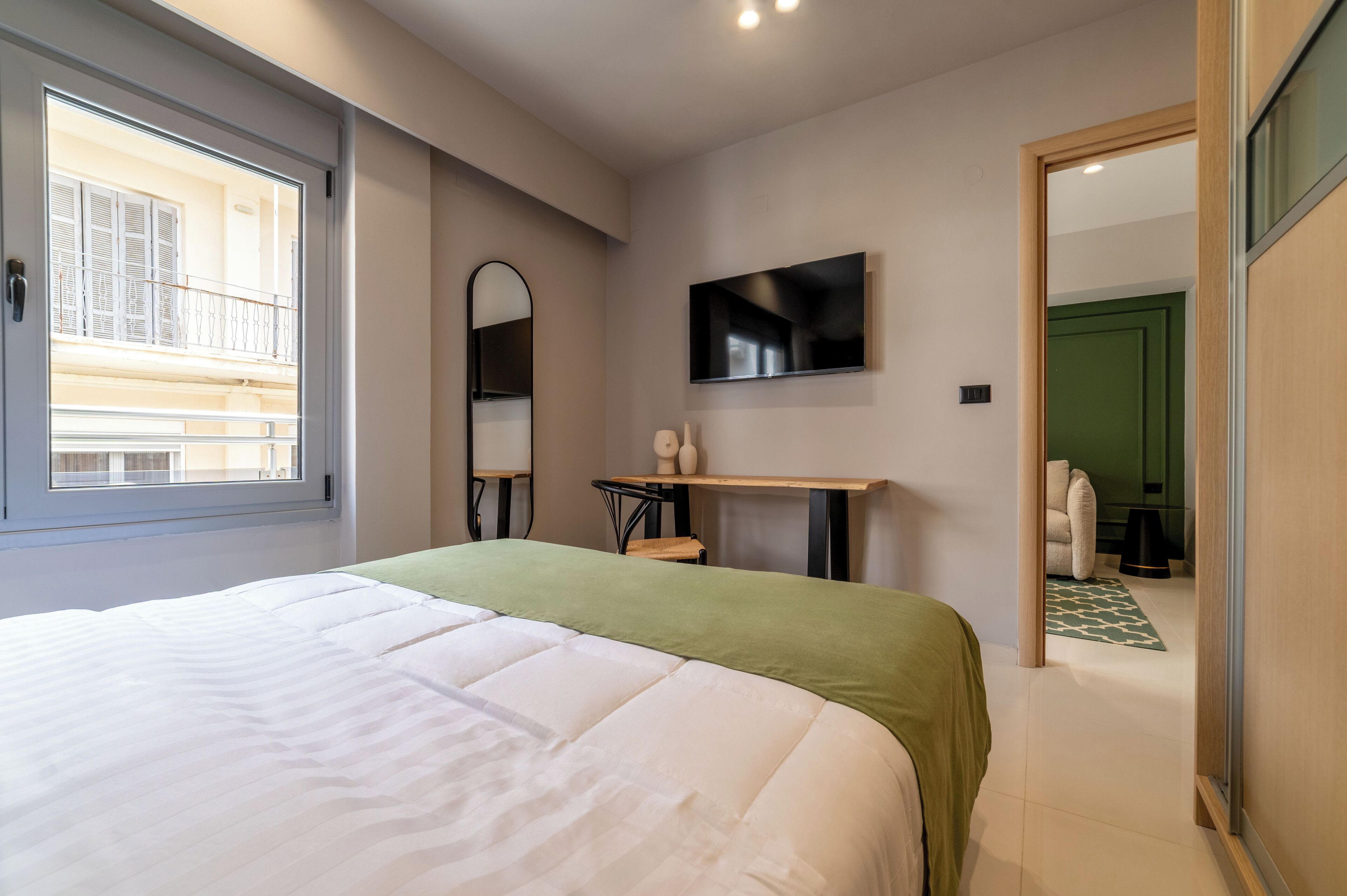 A bedroom with a large bed, a TV mounted on the wall, a mirror, and a window with a view of a balcony.