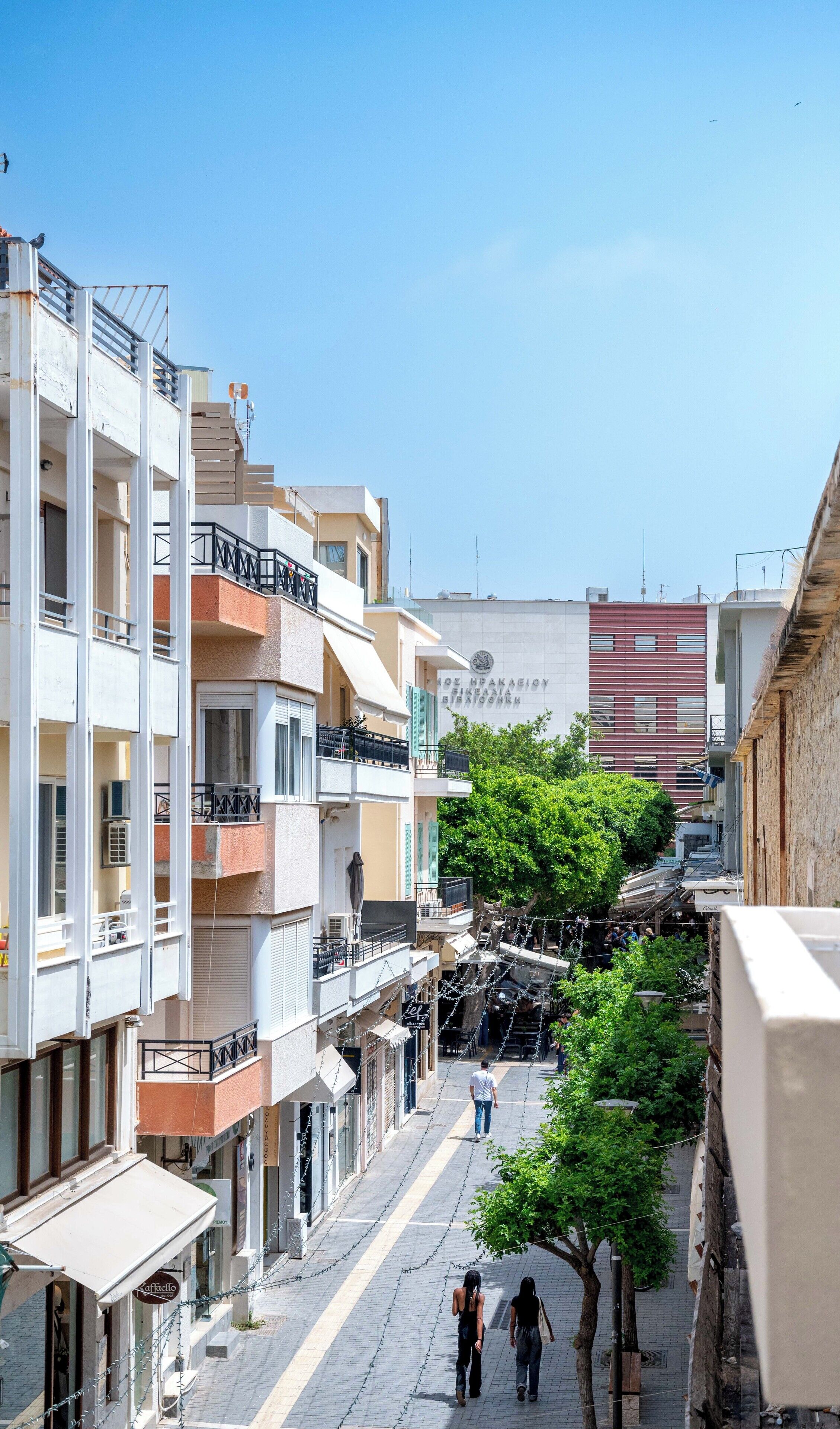 A narrow urban street with multi-story buildings, balconies, and a clear blue sky.