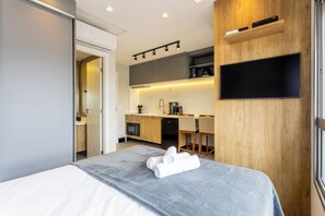 A modern hotel room with a bed, a kitchenette, a flat-screen TV, and a wooden accent wall. - Modern Apartments With Fitness Area and Coworking in the Best Location of Pompeia, SP - DIO (São Paulo)