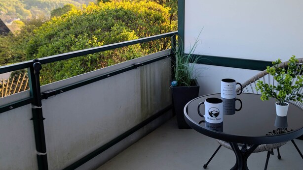 Un balcón con una silla de mimbre, una mesita con una taza y una planta en maceta.