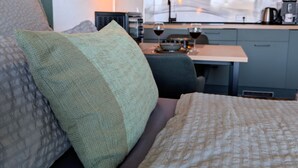 Un dormitorio con cama, almohadas, una mesita de noche y vistas a una zona de cocina con armarios y un ventanal.