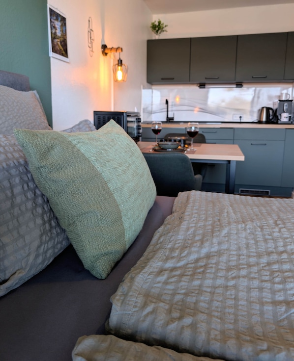 A bedroom with a bed, pillows, a nightstand, and a view of a kitchen area with cabinets and a window. - cozy MeLou Fewo Harzruhe Wellness (Bad Lauterberg)