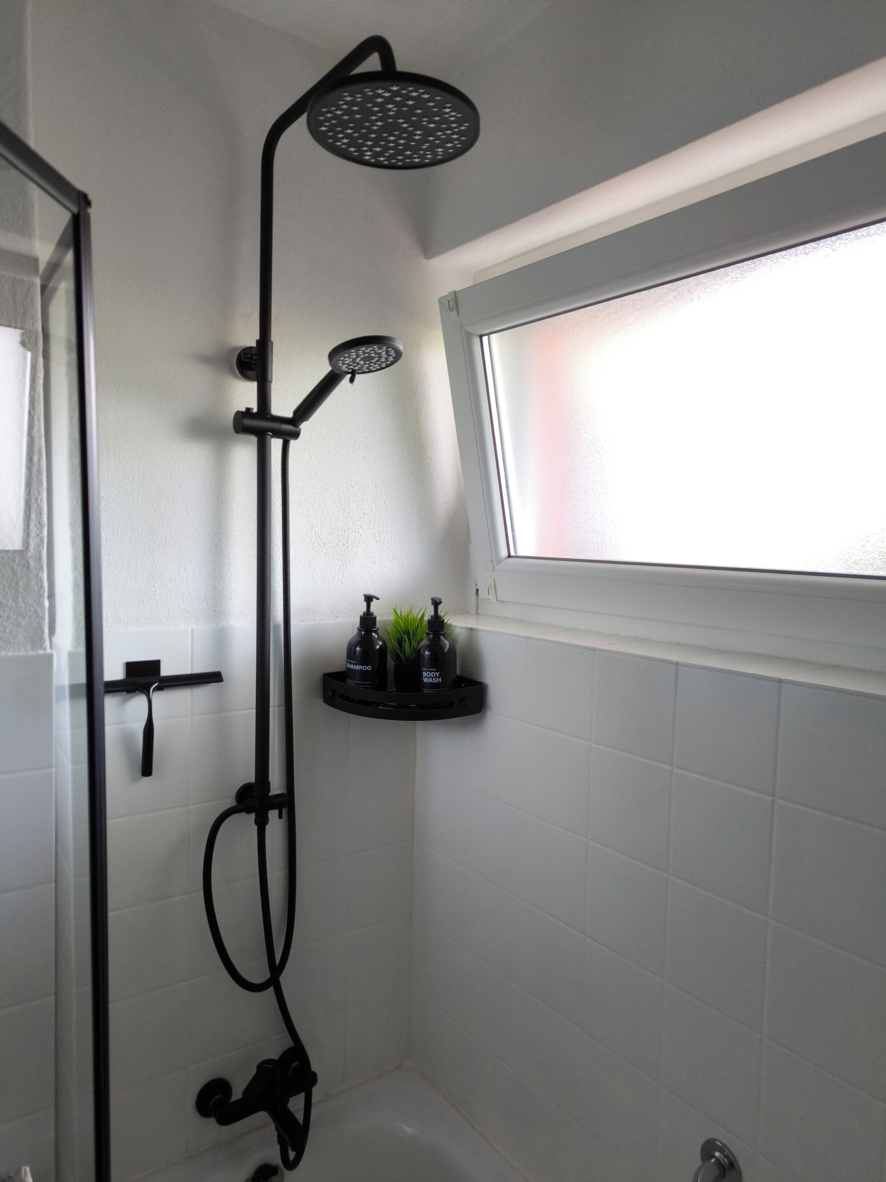 Ducha con cabezal de ducha de mano y cabezal de ducha tipo lluvia, una ventana sobre la ducha y un estante con botellas de champú y gel de baño.