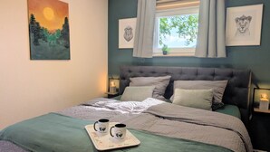 A bedroom with a bed, a window with curtains, a painting on the wall, and a tray with mugs.