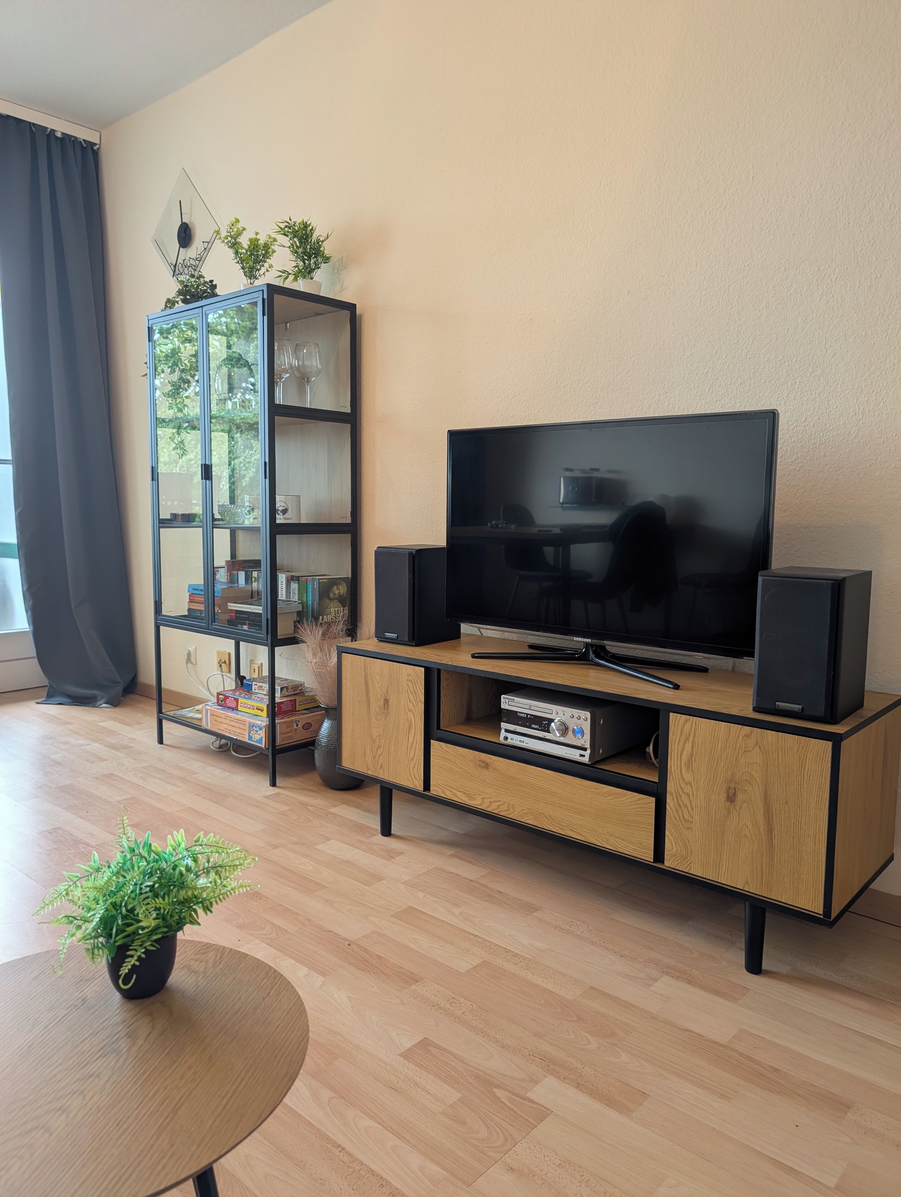 A modern living room with a flat-screen TV, wooden entertainment unit, and a glass-fronted shelving unit with decorative items.