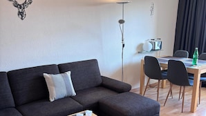 A modern living room with a sectional sofa, a small wooden table, a red rug, a dining area with a table and chairs, and a wall-mounted lamp.