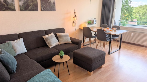 A modern living room with a sofa, ottoman, and a dining table with chairs. There are two wall-mounted paintings, a lamp, and a window with curtains.