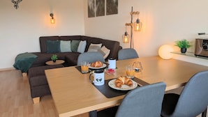 A modern living room with a wooden dining table, grey upholstered sofa, and a coffee table with breakfast items.