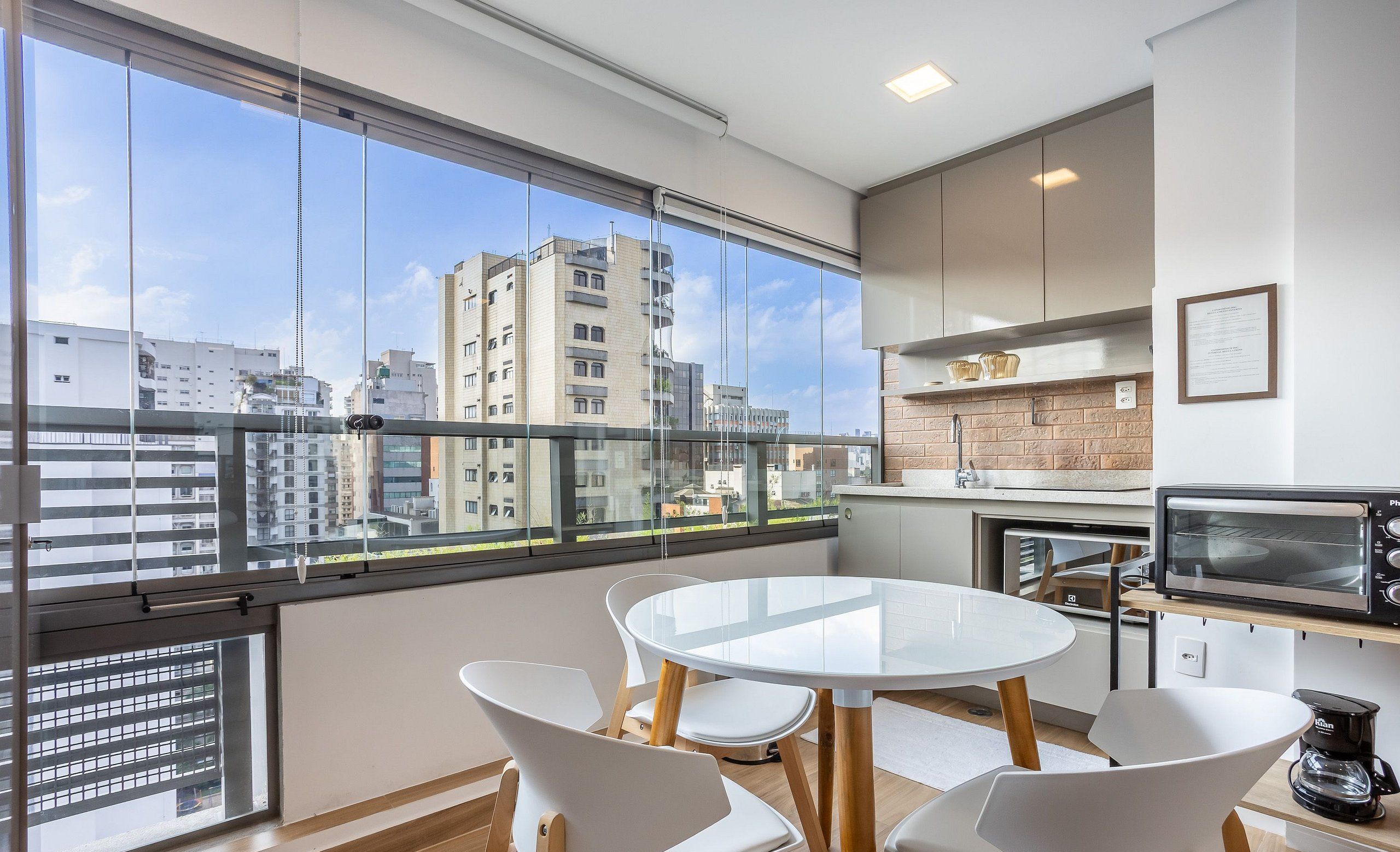 Uma cozinha moderna com uma mesa de jantar redonda e cadeiras brancas, amplas janelas com vista para a cidade e forno embutido.