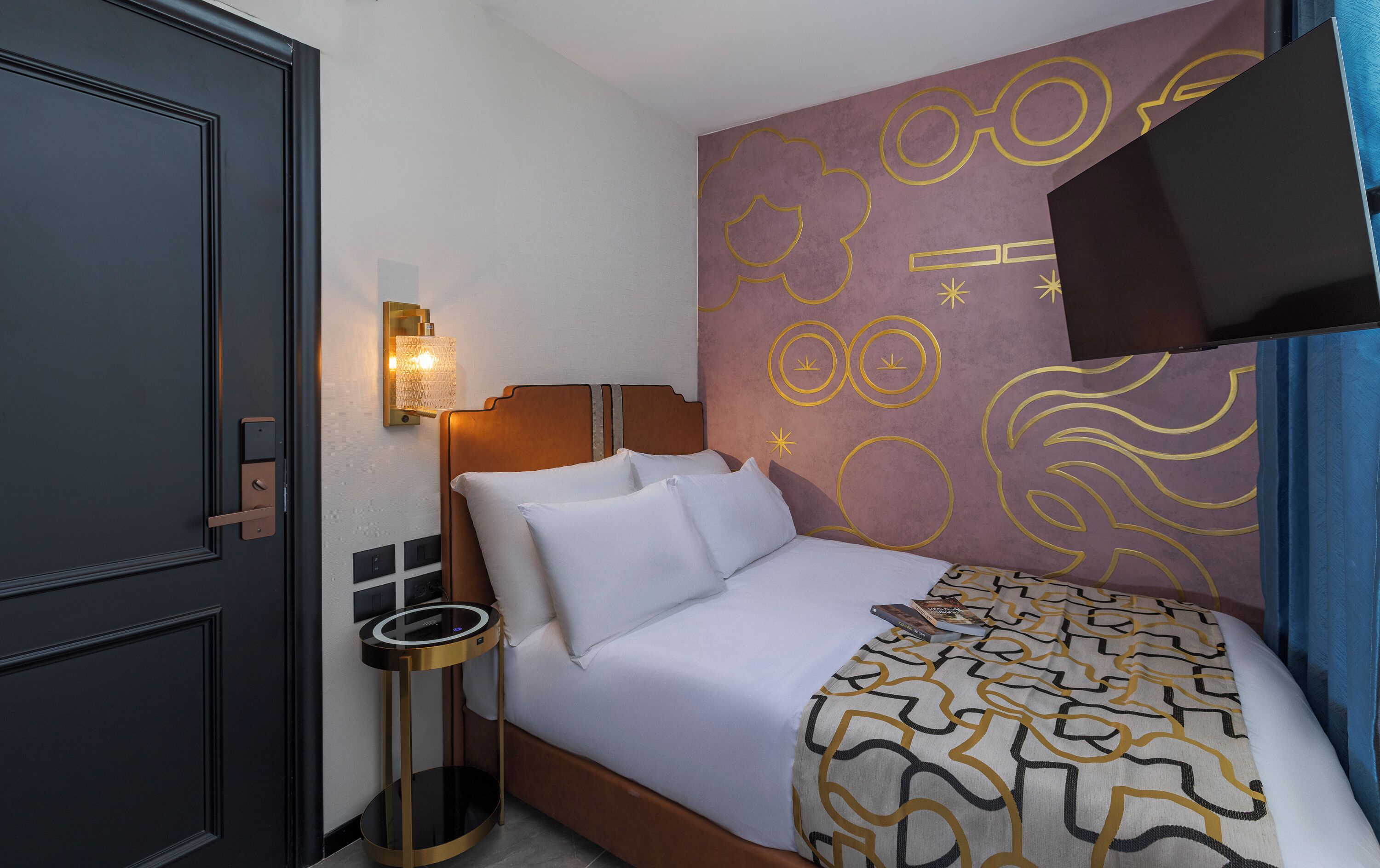 Una habitación de hotel con una cama, una mesita de noche, una lámpara de pared y un televisor.